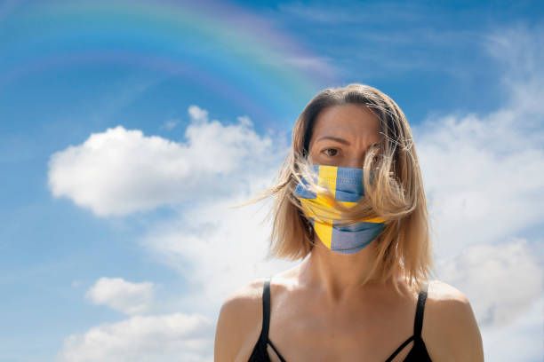 istock Woman is Looking into Camera While Wearing Swedish Flag Protective Mask To Avoid Coronavirus Covid-19 Against Cloudscape with Rainbow 1251690193