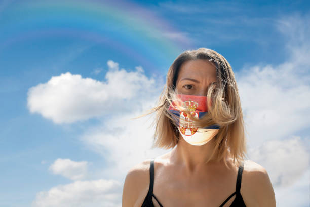 istock Woman is Looking into Camera While Wearing Serbian Flag Protective Mask To Avoid Coronavirus Covid-19 Against Cloudscape with Rainbow 1251690471