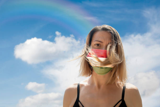 istock Woman is Looking into Camera While Wearing Hungarian Flag Protective Mask To Avoid Coronavirus Covid-19 Against Cloudscape with Rainbow 1251690632