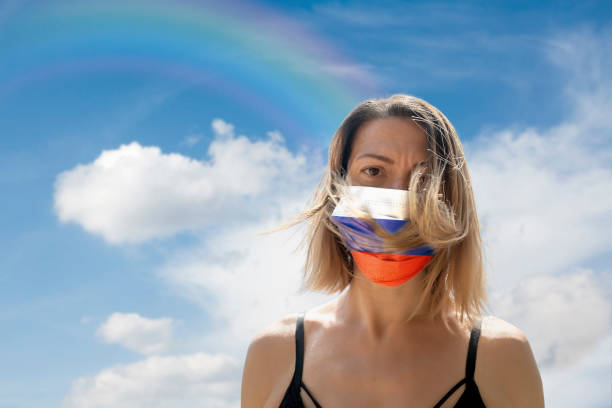 istock Woman is Looking into Camera While Wearing Russian Flag Protective Mask To Avoid Coronavirus Covid-19 Against Cloudscape with Rainbow 1251690980