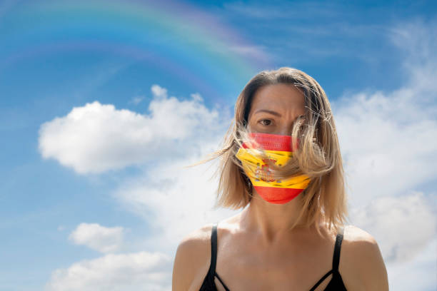 istock Woman is Looking into Camera While Wearing Spanish Flag Protective Mask To Avoid Coronavirus Covid-19 Against Cloudscape with Rainbow 1251691029