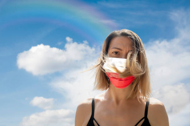 istock Woman is Looking into Camera While Wearing Polish Flag Protective Mask To Avoid Coronavirus Covid-19 Against Cloudscape with Rainbow 1251691082