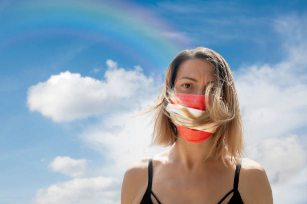 istock Woman is Looking into Camera While Wearing Austrian Flag Protective Mask To Avoid Coronavirus Covid-19 Against Cloudscape with Rainbow 1251691772