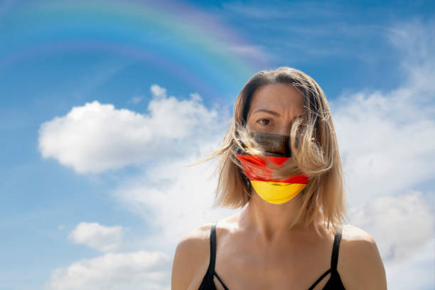 istock Woman is Looking into Camera While Wearing German Flag Protective Mask To Avoid Coronavirus Covid-19 Against Cloudscape with Rainbow 1251691975