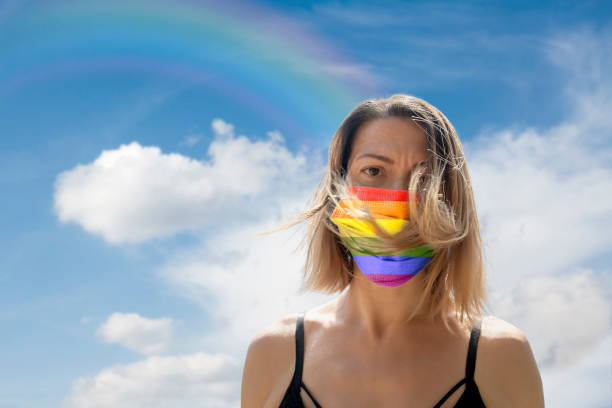 istock Woman is Looking into Camera While Wearing Gay Pride Flag Protective Mask To Avoid Coronavirus Covid-19 Against Cloudscape with Rainbow 1251692215
