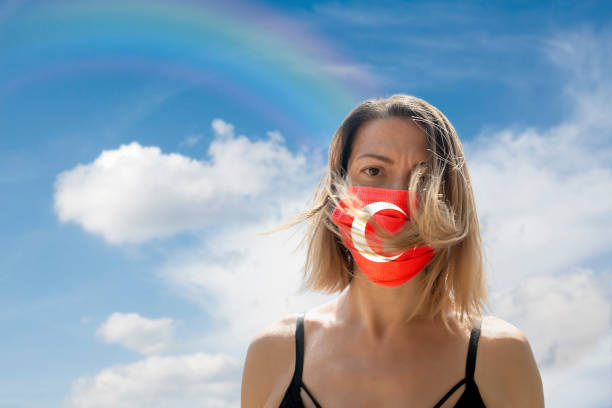 istock Woman is Looking into Camera While Wearing Turkish Flag Protective Mask To Avoid Coronavirus Covid-19 Against Cloudscape with Rainbow 1251692354