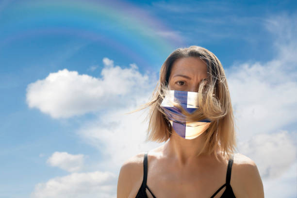 istock Woman is Looking into Camera While Wearing Finnish Flag Protective Mask To Avoid Coronavirus Covid-19 Against Cloudscape with Rainbow 1251692415
