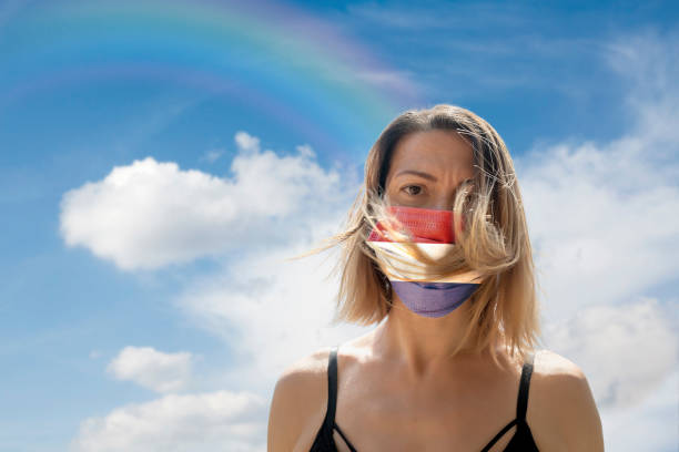istock Woman is Looking into Camera While Wearing Dutch Flag Protective Mask To Avoid Coronavirus Covid-19 Against Cloudscape with Rainbow 1251692514