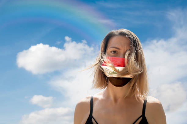 istock Woman is Looking into Camera While Wearing Iraqi Flag Protective Mask To Avoid Coronavirus Covid-19 Against Cloudscape with Rainbow 1251692882