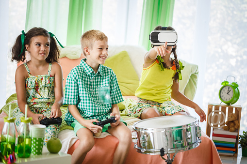 Front view of three kids sitting on the bedroom and enjoying afternoon with instruments and video games of virtual reality.
