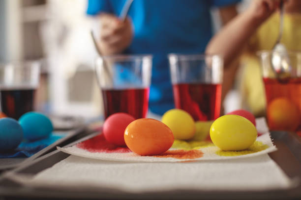 Selective focus of different colored eggs lying on a table while in the blurred background unrecognizable sibling are dying them for Easter.