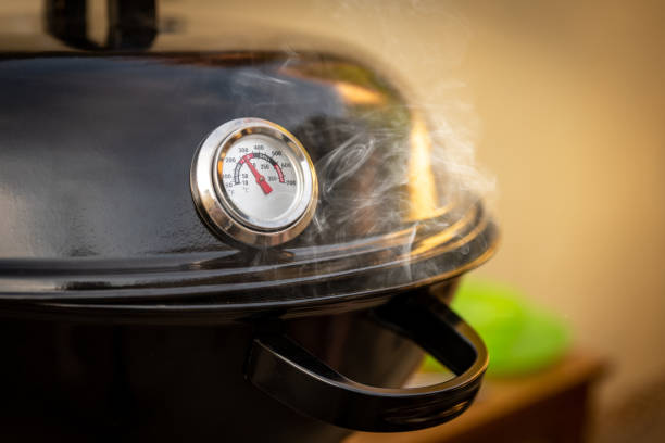 A round thermometer showing 130 degrees Celsius placed on a charcoal grill.
