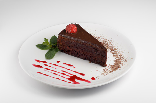 Chocolate cake with a beautiful serving on a white plate decorated with jam, a sprig of mint, a strawberry and chocolate chips. For use in menu design and online delivery service or cooking book
