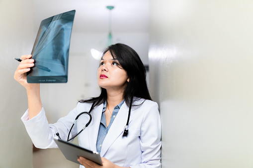 A photo of confident asian female doctor examining x-ray in hospital. Young professional in lab coat is analysing medical report. Expert is standing at brightly lit workplace.