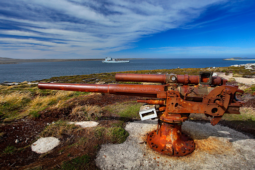 Falklands War, rocky coast with old rusty cannon. Corroded artillery gun from Falklands Conflict in nature habitat. Blue sky landscape, Falkland Islands. Military ship on sea. Memorial war monument.