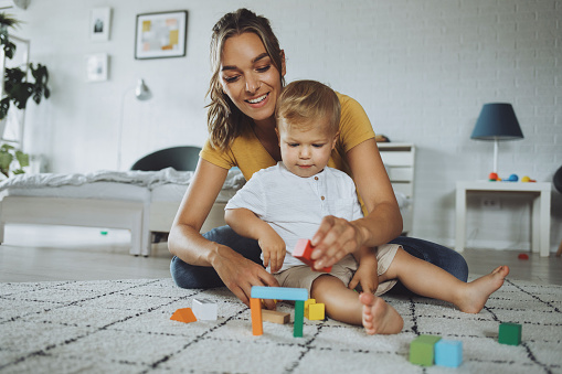 Beautiful young mother and cute baby boy playing with construction toys in their living room.