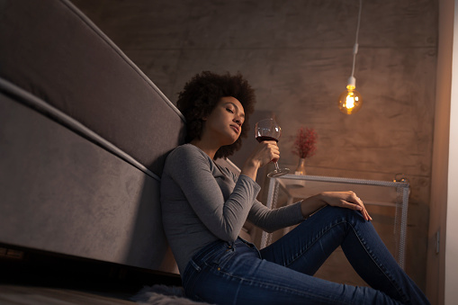 Beautiful young mixed race woman sitting on the floor by the bed, having a glass of wine and relaxing at home after a busy day