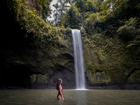 Woman in red bikini in water looking at waterfall. Tibumana waterfalls in bali. This place is very famous.
