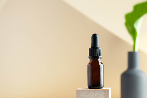 Serum oil for the face in a dark glass flask on a light background, in the background is a green plant with a place to enter text from above. Skin care.