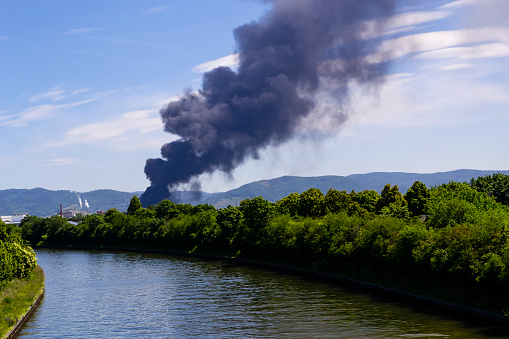 a huge column of black smoke of a fire on a factory rising up to the sky. A landscape with a river in the foreground, a black smoke of a fire in the middle, green hills and blue sky in the background.