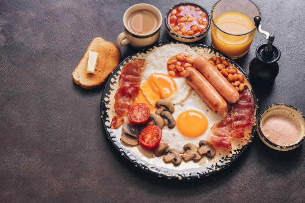 istock English traditional breakfast on a dark brown rustic background, copy space. Fried eggs with bacon, sausages and beans, coffee, orange juice. 1251744120