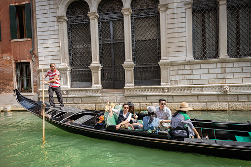 Venice, Veneto, Italy - Sep 13th, 2015: five tourists of Asian ethnicity sitting in the gondola with a gondolier, typical Venetian rowing boat. Sightseeing tour along the Canals of the famous city. The gondola is a traditional, flat-bottomed Venetian rowing boat, well suited to the conditions of the Venetian lagoon. It is similar to a canoe, except it is narrower. It is propelled by a gondolier, who uses a rowing oar, which is not fastened to the hull, in a sculling manner and acts as the rudder.