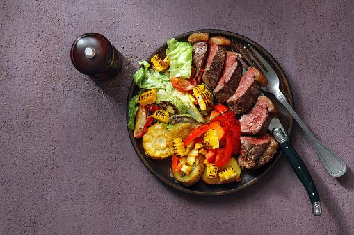Grilled Steak with Potato Salad and Vegetables. Flat lay top-down composition on purple background. Horizontal image with copy space.