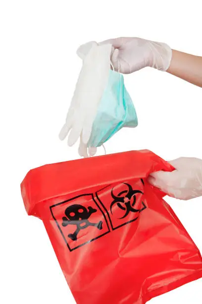 Photo of Dropping used medical mask and medical gloves in red biohazard bag isolated on white background.