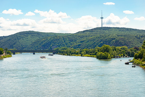 A beautiful view of the River Rhine in West Germany on which ships sail. In the background a dense forest, a visible bridge, a tower and a branch of the river.
