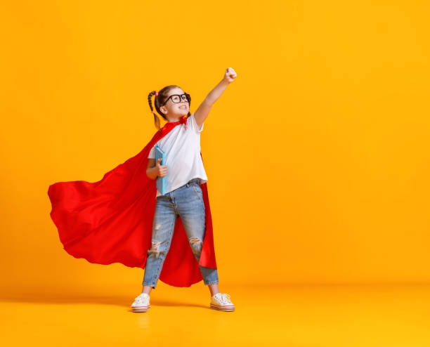Little superhero studying at school Full body girl in superhero cape smiling and raising fist up while being ready for school studies against yellow backdrop cape garment stock pictures, royalty-free photos & images