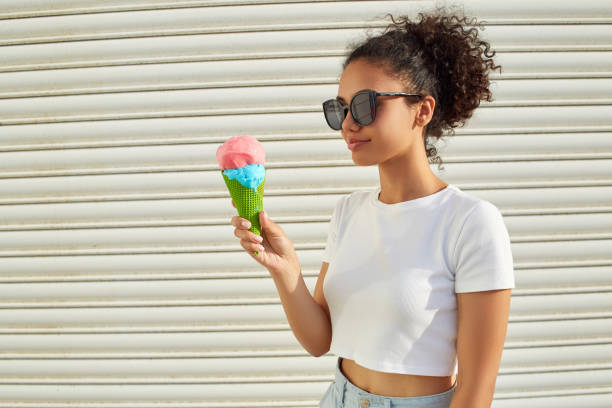 白いtシャツと軽いジーンズを着た若い美しいアフリカ系アメリカ人の女の子は、晴れた日に明るい壁にアイスクリームを食べます。 - women eating ice cream indigenous culture ストックフォトと画像