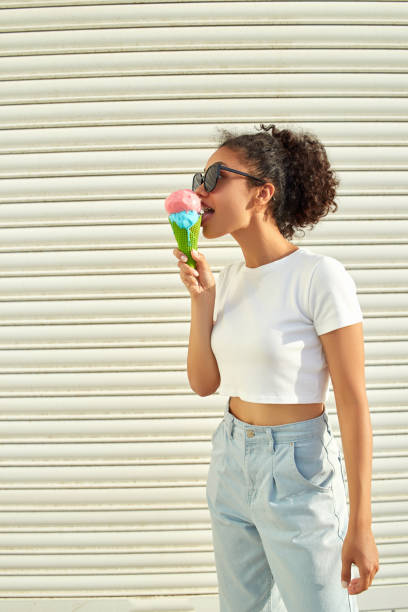 白いtシャツと軽いジーンズを着た若い美しいアフリカ系アメリカ人の女の子は、晴れた日に明るい壁にアイスクリームを食べます。 - women eating ice cream indigenous culture ストックフォトと画像