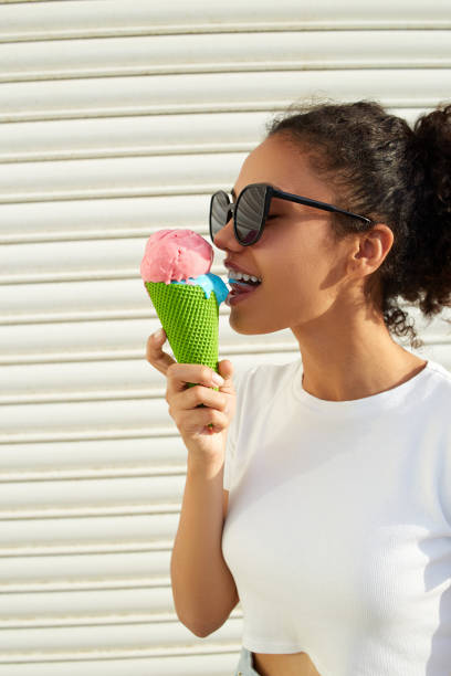 白いtシャツと軽いジーンズを着た若い美しいアフリカ系アメリカ人の女の子は、晴れた日に明るい壁にアイスクリームを食べます。 - women eating ice cream indigenous culture ストックフォトと画像