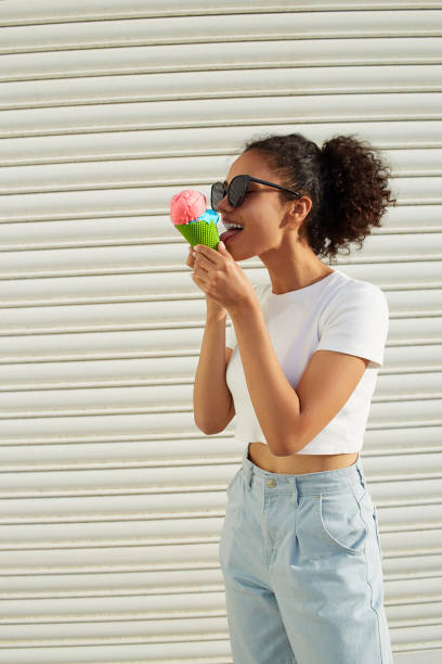 白いtシャツと軽いジーンズを着た若い美しいアフリカ系アメリカ人の女の子は、晴れた日に明るい壁にアイスクリームを食べます。 - women eating ice cream indigenous culture ストックフォトと画像