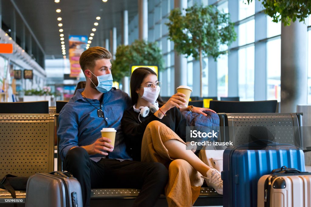 Young couple wearing N95 face masks waiting in airport area Young woman and man traveling by plane during COVID 19, wearing N95 face masks, sitting on bench with take away coffee in airport waiting area. Travel Stock Photo