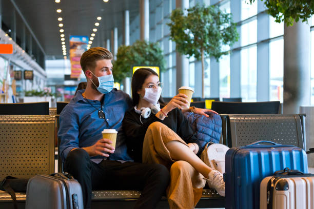 Young couple wearing N95 face masks waiting in airport area Young woman and man traveling by plane during COVID 19, wearing N95 face masks, sitting on bench with take away coffee in airport waiting area. cancellation photos stock pictures, royalty-free photos & images
