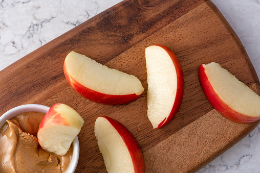 Five apple wedges on wooden cutting board with a small bowl of peanut butter for dipping. One apply wedge is in the peanut butter, healthy snack. Shot from above, white granite countertop.