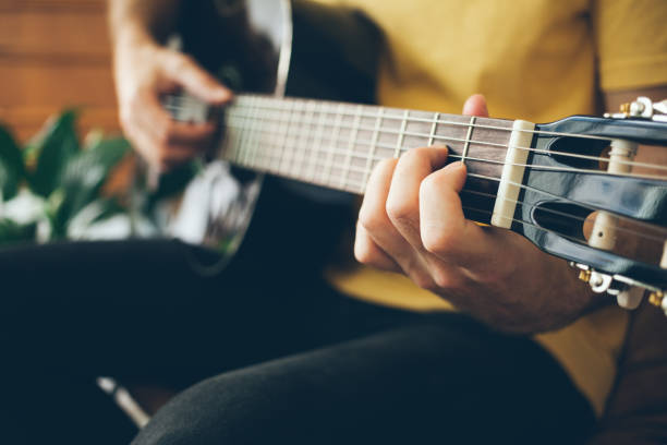 Close-up of a man hands playing black color guitar at home in beautiful modern interior. Natural light and selective focus. Guitarist in yellow t-shirt. chord stock pictures, royalty-free photos & images
