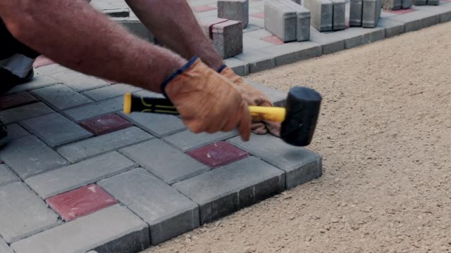 A gloved craftsman is laying concrete tiles using a bubble level and a rubber mallet. Tile laying sounds. Brick walkway paved with professional paver. Laying gray concrete paving slabs in the courtyard of the house on gravelly sandy base