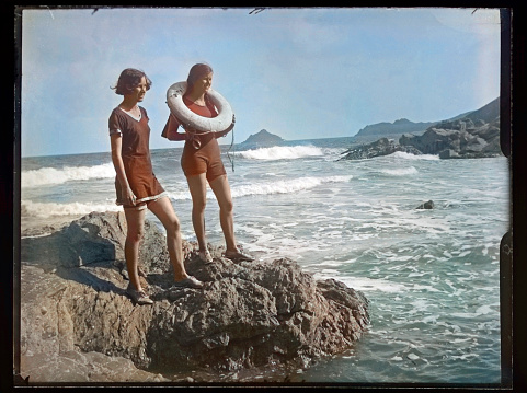 Colourised antique photograph from Edwardian era of two teenage girls at the seaside, one with a Lifebuoy round her neck. circa 1910.