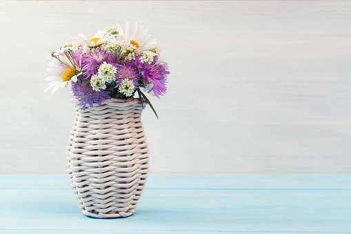 Delicate cute wildflowers in a light wicker vase on a blue wooden table, copy space for text.