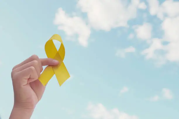 Photo of Hands holding yellow gold ribbon over blue sky, Sarcoma Awareness, Bone cancer, childhood cancer awareness, September yellow, World Suicide Prevention Day concept