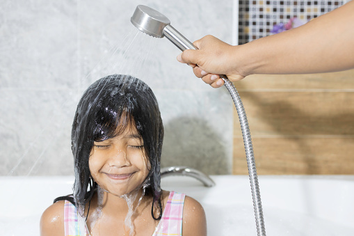 An Asian girl is washing her hair in the bathroom, with her mother helping to spray water from the shower to clean her hair.