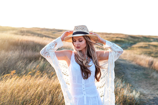 Beautiful woman wearing in white dress and straw hat enjoying nature outdoors at sunset.