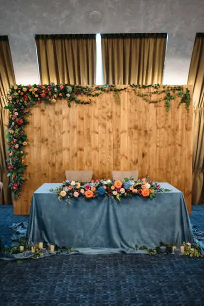 Wedding presidium in restaurant, free space. Wedding banquet table for newlyweds with flowers, greenery, blue velvet and candles . Lush floral arrangement on wedding table