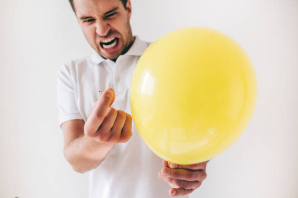 istock Young man isolated over white background. Guy emotional expressing before piecercing a yellow balloon with needle. Cut view. 1252332011