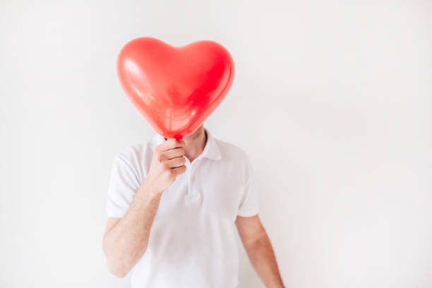 istock Young man isolated over white background. Guy hold red balloon in heart shape in front face. Cover or hide it. Love. 1252332014