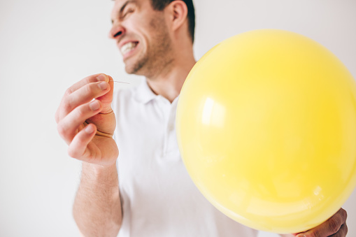 Young man isolated over white background. Guy pierce a balloon with needle. Afraid of sound. Cut view and close up. Defocused guy on picture