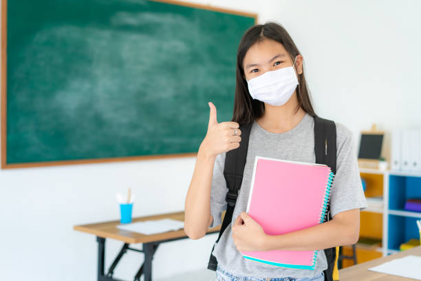 アジアの小学生の女の子の親指を立て、学校に戻っている間に教室でcovid 19の発生を防ぐためにマスクを着用し、教育の概念のための新しい正常。 - thumbs up book smiling student ストックフォトと画像