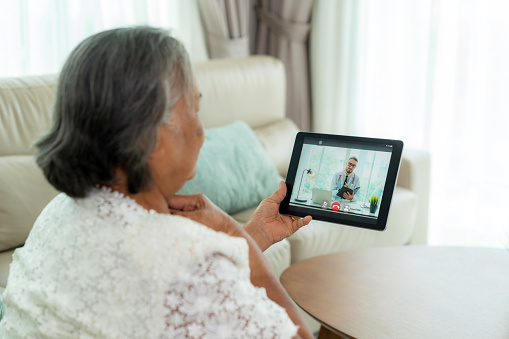 Back view of elderly woman making video call with her doctor with her feeling sore throat on digital tablet online healthcare digital technology service consultation while staying at home.